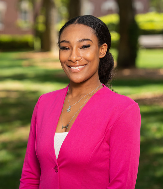 A smiling young woman wearing a pink blazer, standing outdoors in a sunny park setting.