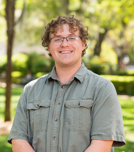 A young man with curly hair and glasses smiles confidently in a green park setting.
