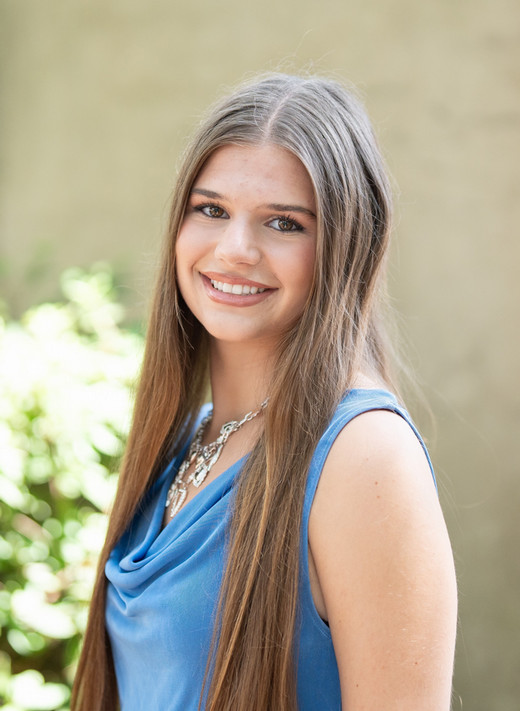 A smiling young woman with long hair wearing a blue top stands outdoors, with greenery in the background.