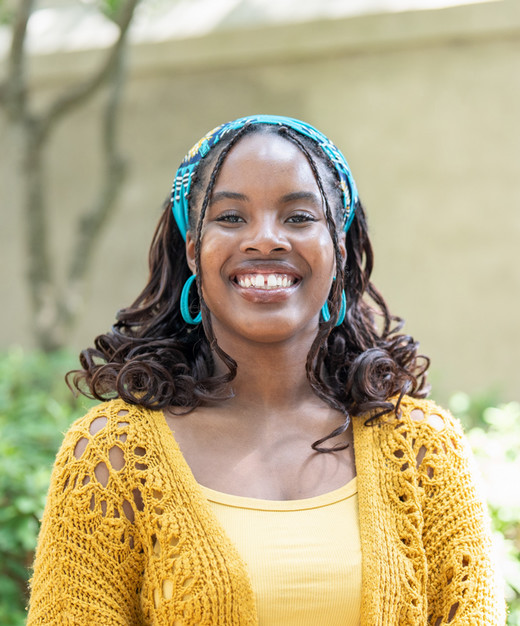 A smiling woman wearing a yellow cardigan and turquoise accessories stands outdoors in a lush green setting.