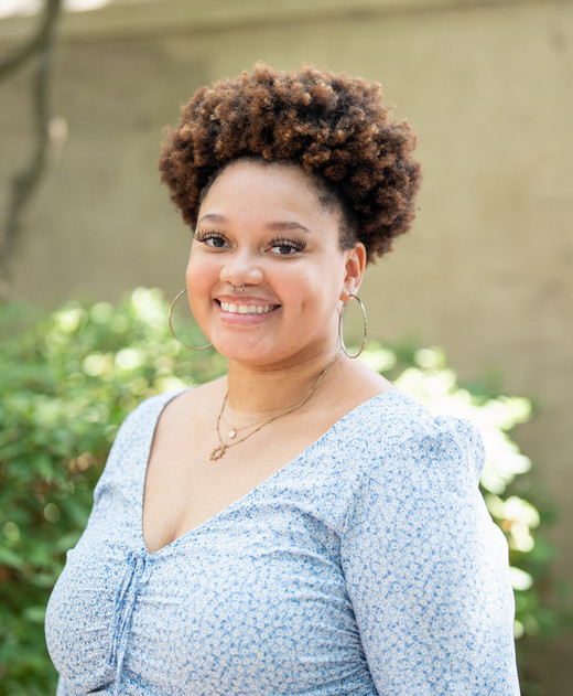 A smiling young woman with curly hair stands outdoors, wearing a light blue floral top and gold hoop earrings.