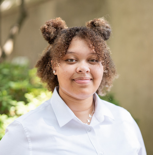 A young woman with curly hair styled in two buns, wearing a white shirt, poses against a greenery backdrop.