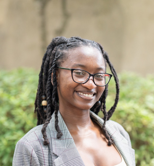 A smiling woman with dreadlocks wearing a gray blazer in front of a green background.