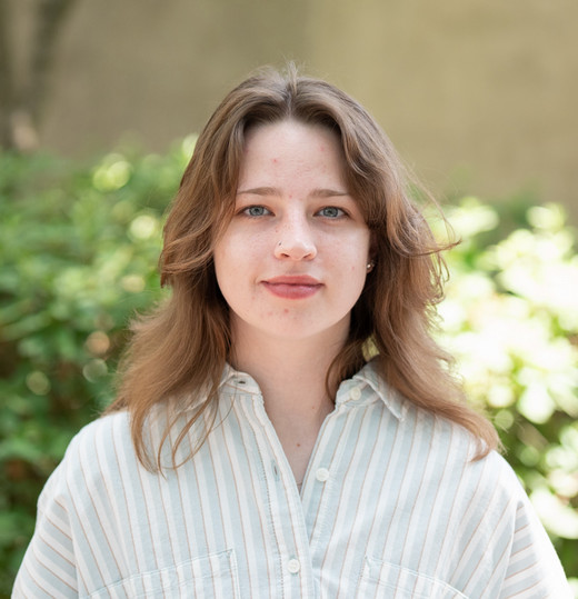 A young woman with shoulder-length wavy hair smiles at the camera, standing in front of greenery.
