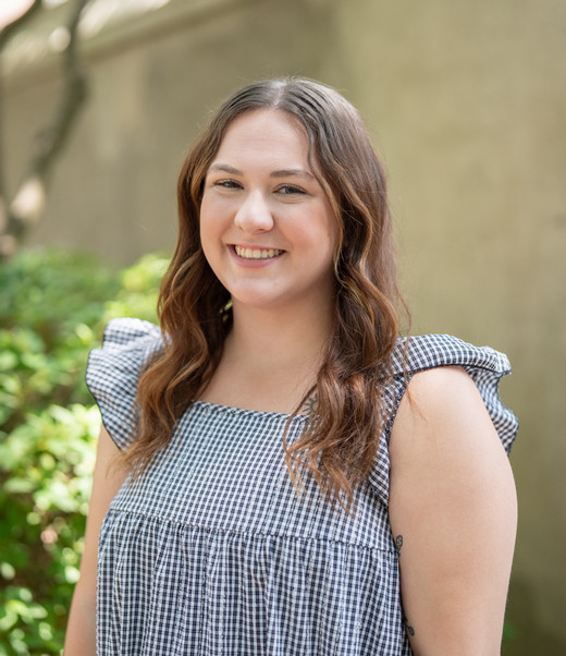 A smiling woman with long wavy hair wearing a gingham-patterned dress stands outdoors surrounded by greenery.
