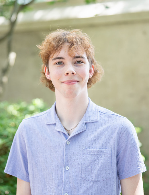 A young man with curly hair smiles confidently while wearing a light purple button-up shirt in a natural outdoor setting.