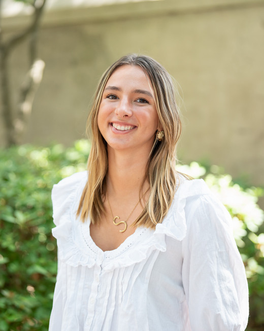 A smiling woman with long, wavy hair wearing a white blouse, standing in front of greenery.