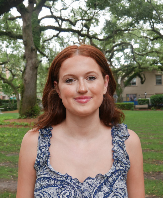 A smiling young woman with wavy red hair stands in a park, surrounded by trees.