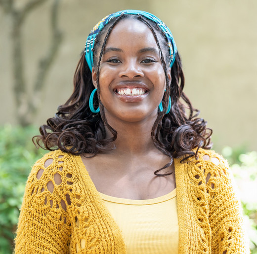 A smiling woman wearing a yellow cardigan and turquoise accessories stands outdoors in a lush green setting.