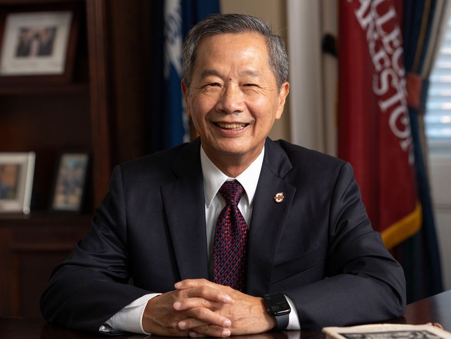 Professional portrait of College of Charleston President Andrew T. Hsu, smiling sitting at a desk with his hands folded in front of him.