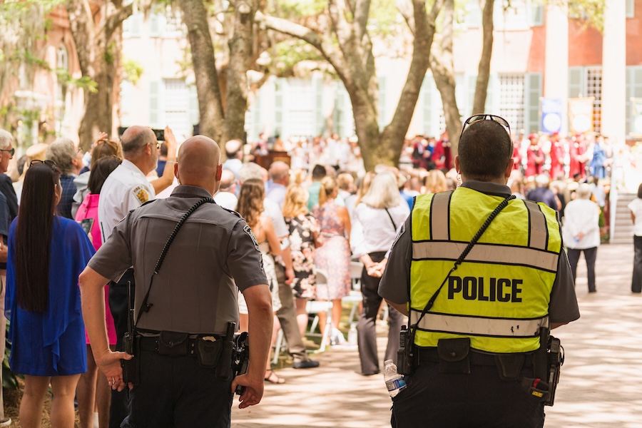 Two police officers stand at the edge of a crowd gathered outdoors, facing a public event in the distance.