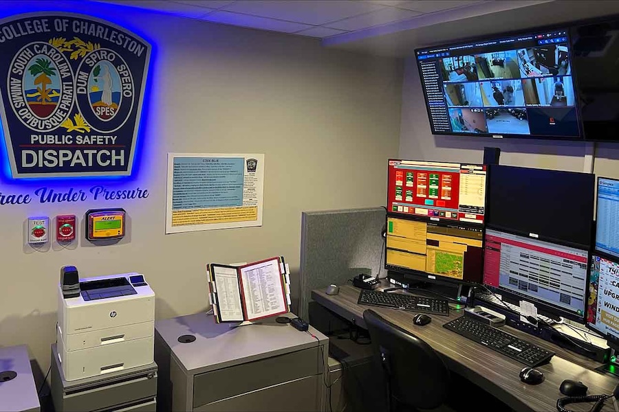 Public safety dispatch office with multiple computer monitors showing call and surveillance screens, plus a wall-mounted “College of Charleston Public Safety Dispatch” sign.