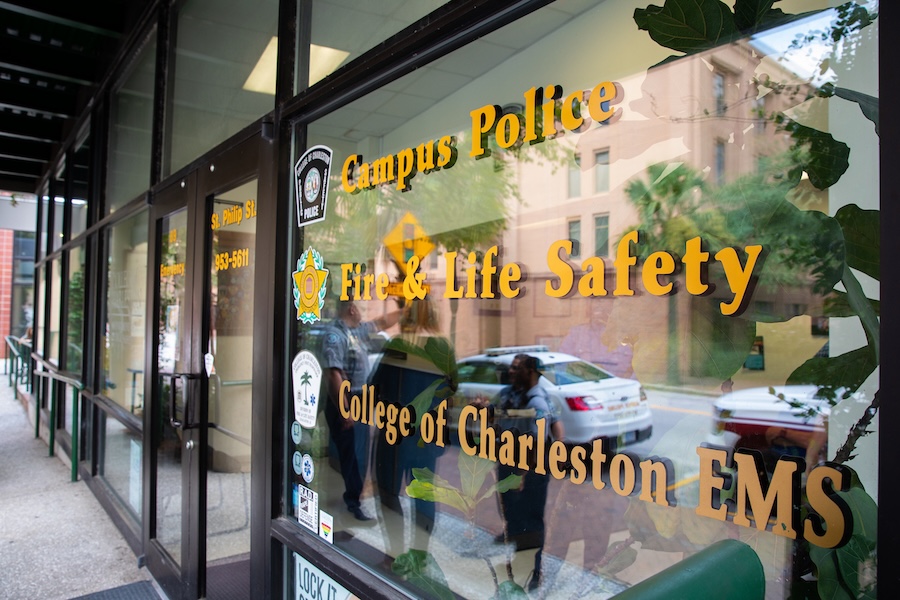 Glass entrance door with gold lettering for Campus Police, Fire & Life Safety, and College of Charleston EMS, reflecting nearby street and buildings.