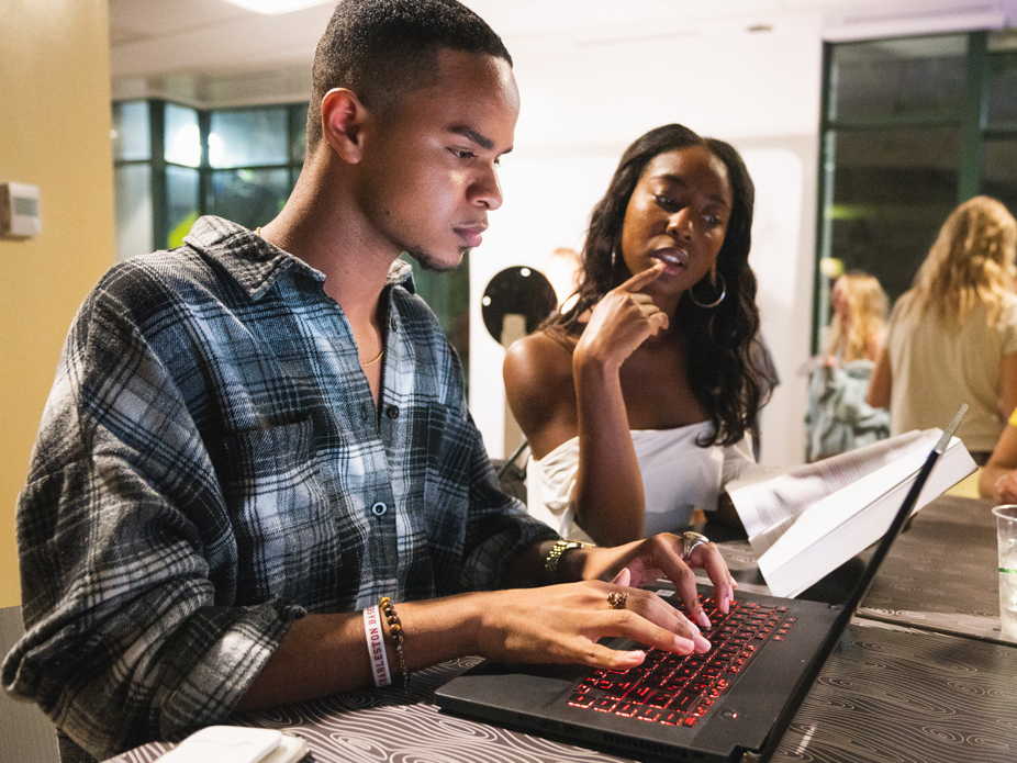 Two students collaborate at a desk, with one typing on a laptop while the other reviews printed documents.