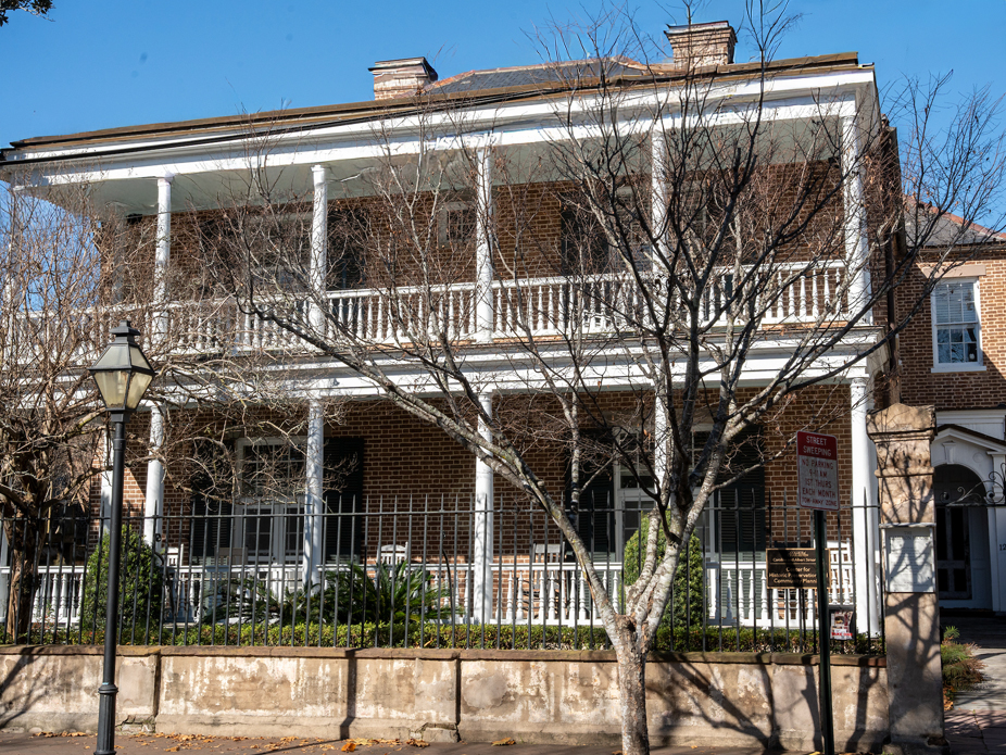 Historic brick building with a large porch and columns, featuring bare trees in front.