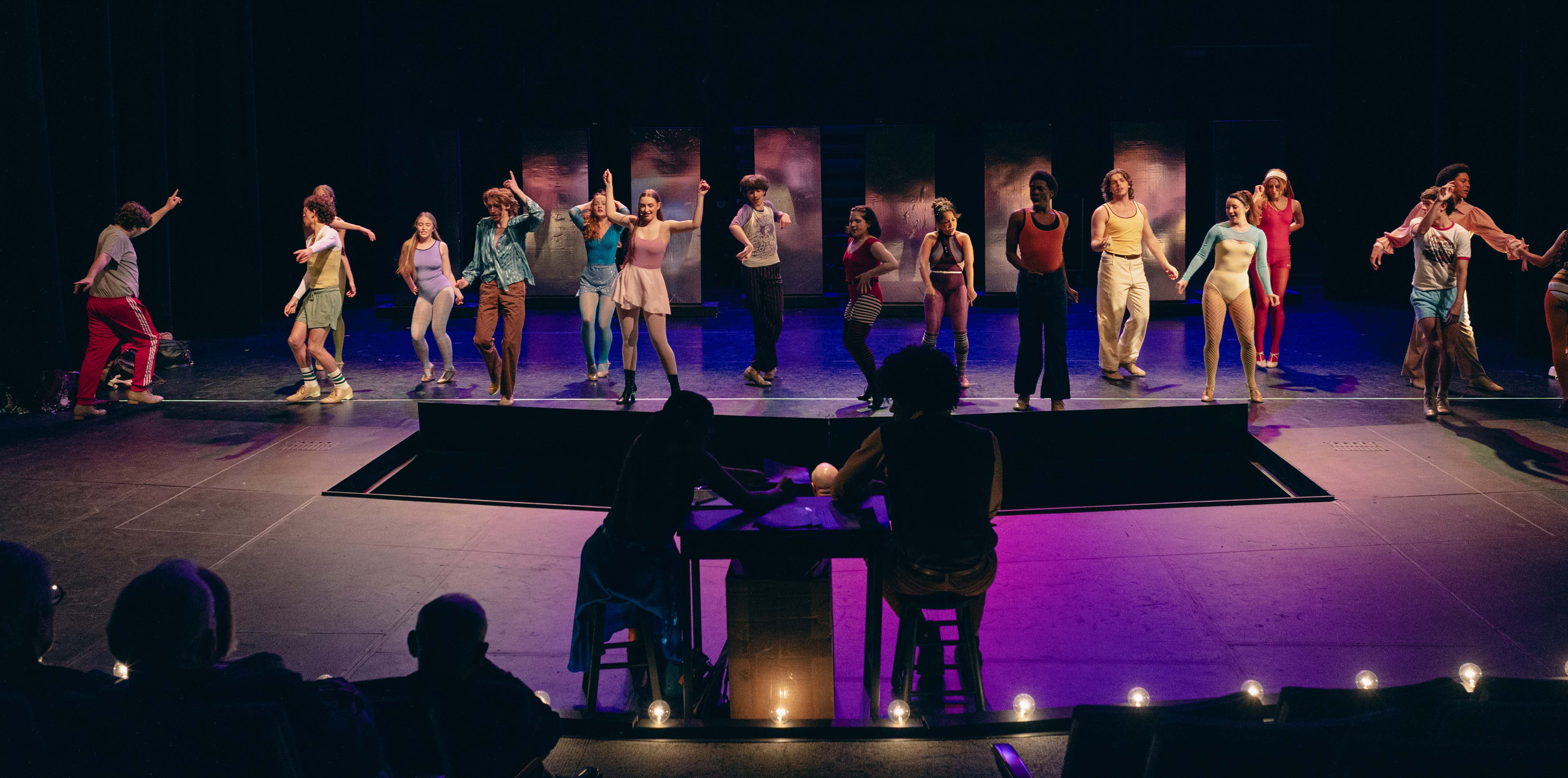 Ensemble of performers in The Chorus Line in colorful retro dance outfits spread across a stage under purple and blue lighting.