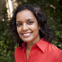 Portrait of Nandini McCauley, a woman with curly hair wearing a red shirt, smiling in a natural outdoor setting.