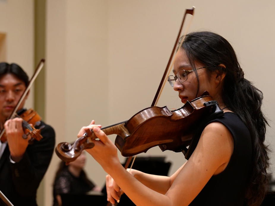 A female violinist performs with other members of the orchestra in the background. 