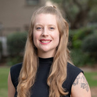 Portrait of Emma Sheehan, a smiling woman with long hair and a tattoo, wearing a black sleeveless top, stands outdoors with greenery in the background.