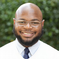 Professional portrait of a Noah Thomas, smiling man with glasses, a beard, and a tie, set against a blurred green background.
