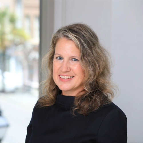 Portrait of a woman with wavy blonde hair, wearing a black top, smiling in a well-lit indoor setting.