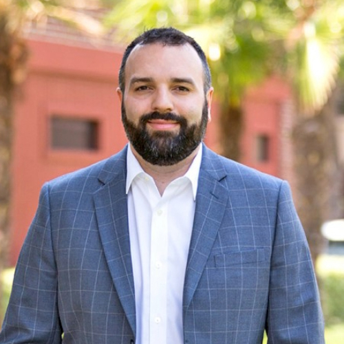 A professionally dressed man with a beard stands outdoors against a backdrop of palmetto trees.
