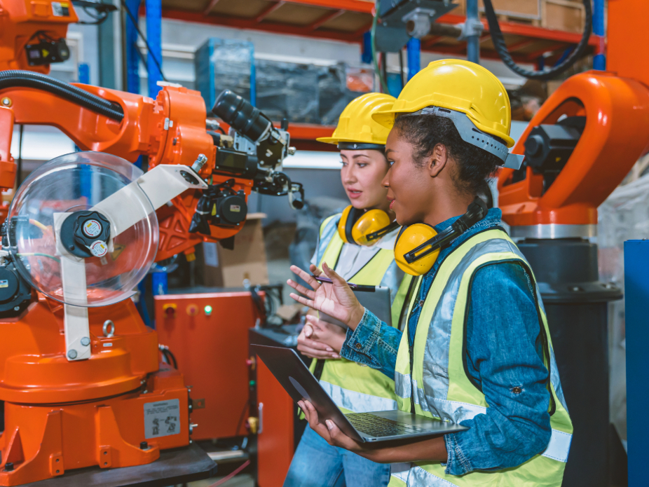 Two women wearing hard hats and safety vests discuss operational details in front of an orange industrial robotic arm in a manufacturing setting.