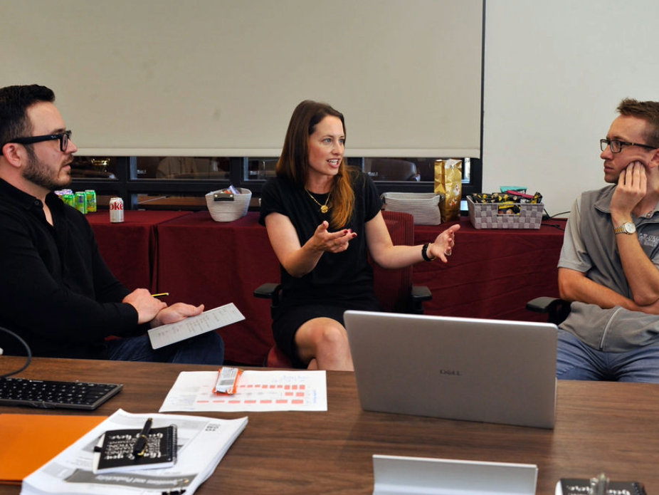 A woman in a black dress engages in discussion with two men in a meeting setting.