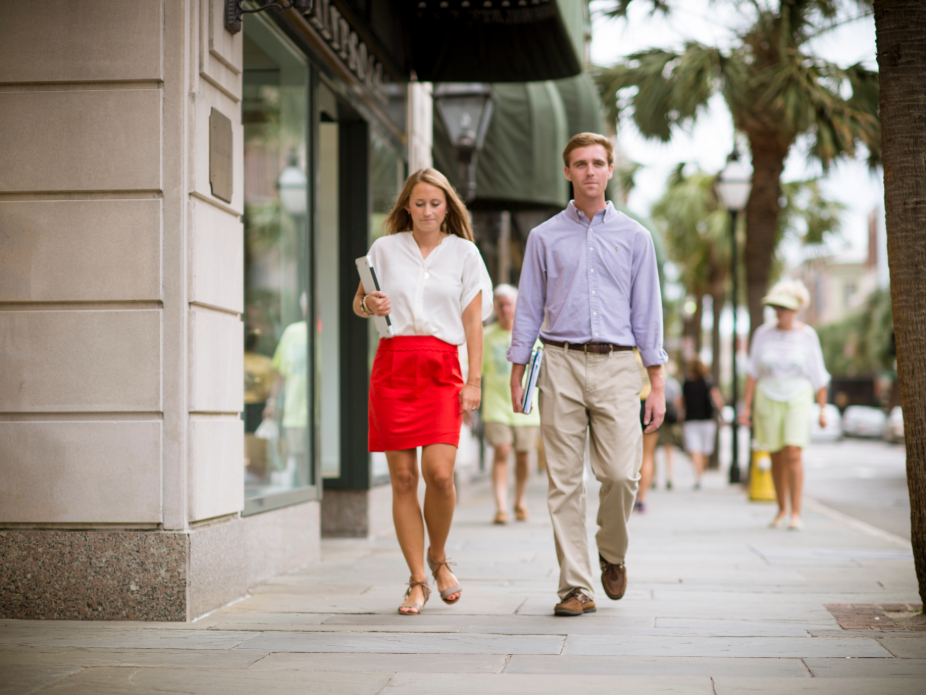 Two young adults walk side by side on a city sidewalk, dressed in smart-casual attire, with palm trees in the background.