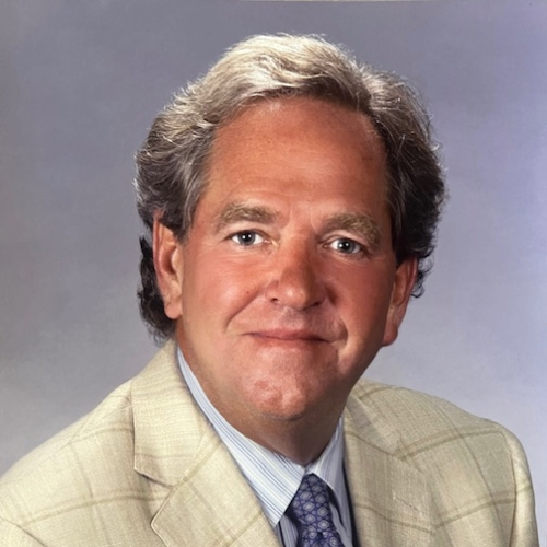 Studio headshot of a middle-aged man with gray hair wearing a light suit jacket and tie against a gray background.