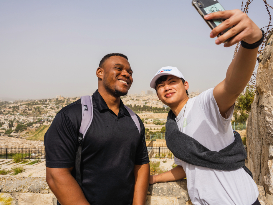 Two people outdoors taking a selfie with a smartphone, standing near a stone wall overlooking a cityscape with historic buildings and hills in the background on a sunny day