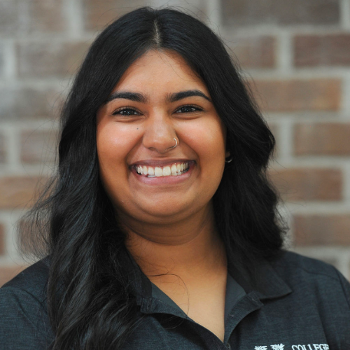 Smiling woman with long black hair wearing a dark grey college shirt against a brick wall.