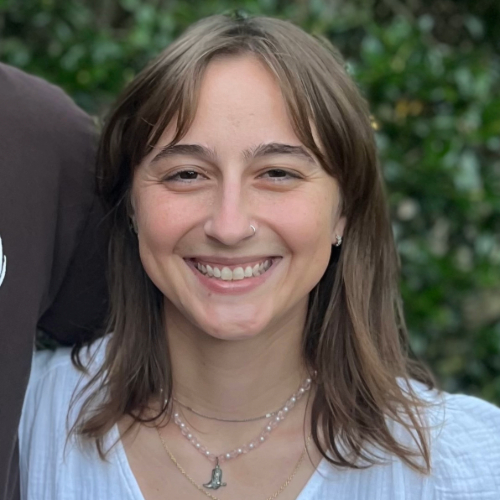 Smiling person with shoulder-length brown hair and a nose ring, wearing layered necklaces against a leafy outdoor background.