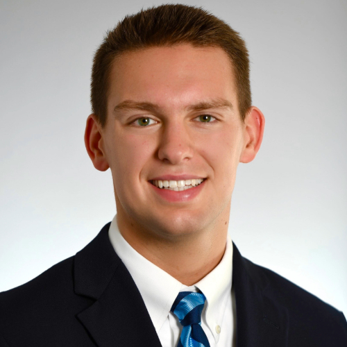 Professional headshot of a young man in a suit and tie, smiling warmly at the camera.