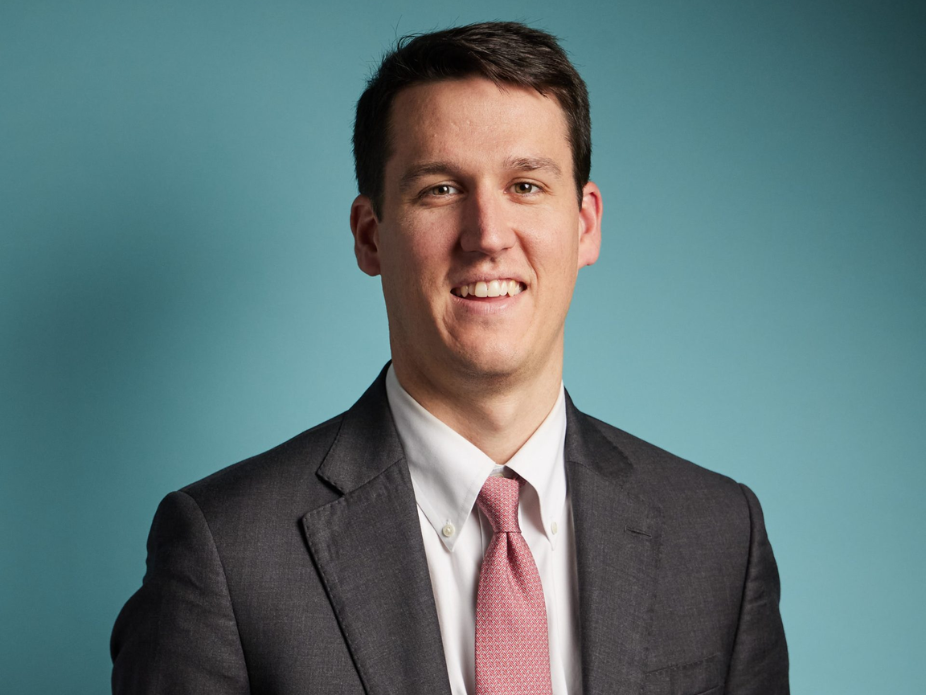 Person wearing a dark suit jacket, white dress shirt, and pink tie, posed against a solid teal background.