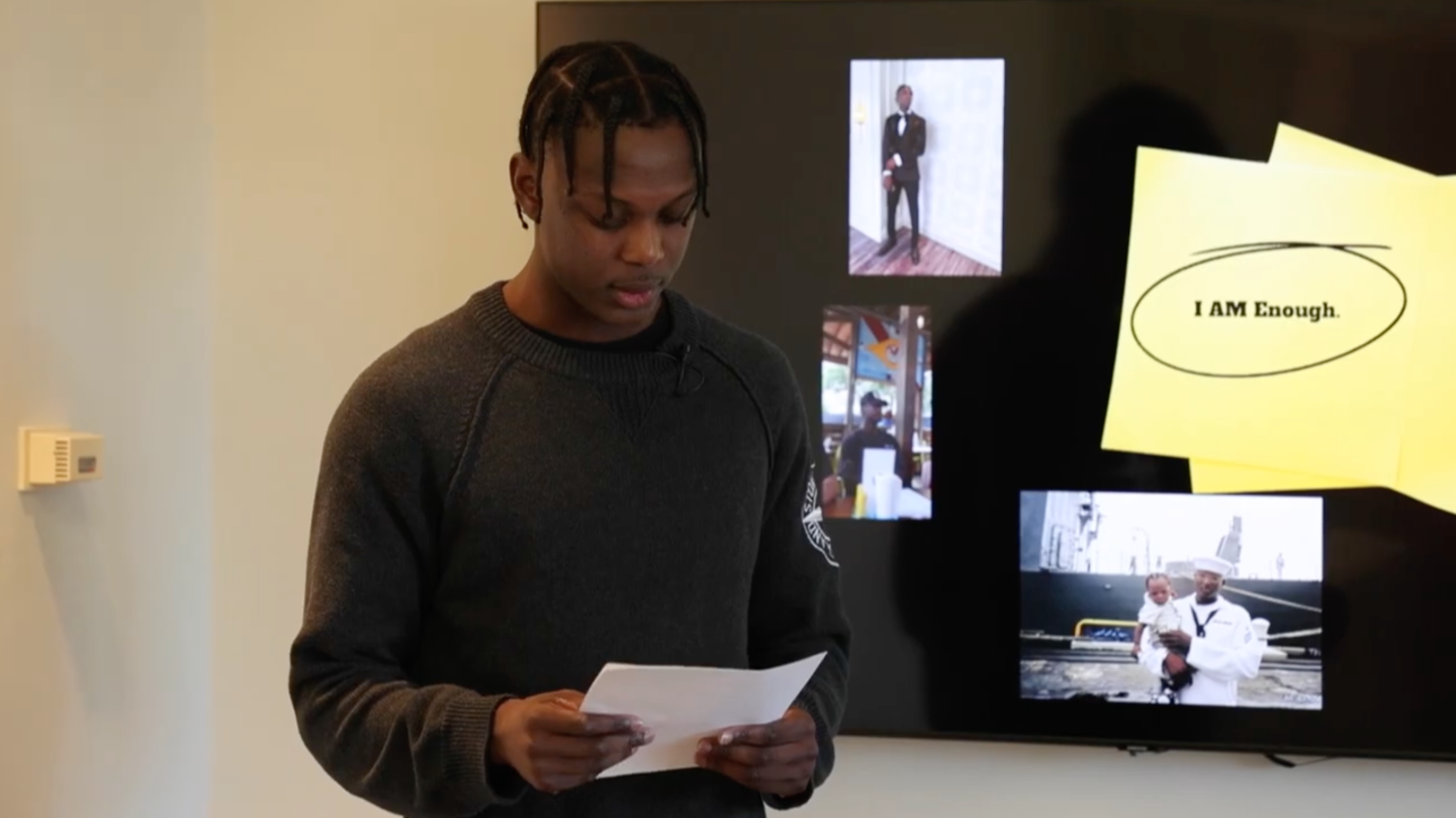 A screenshot of video of young man presenting a class project in a boardroom with a presentation loaded in the background.