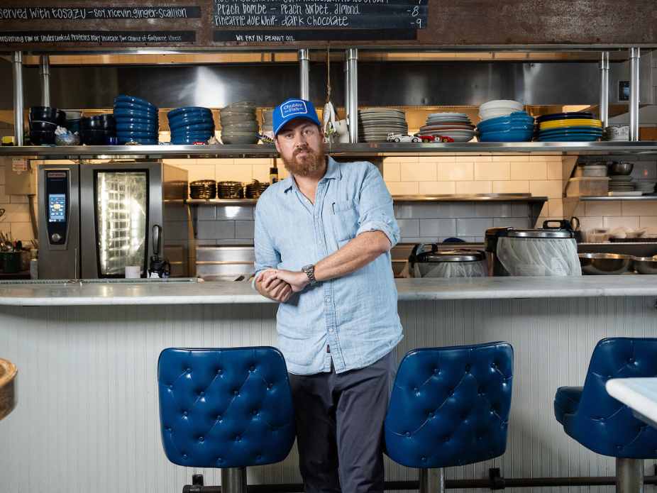 Person standing in front of a restaurant counter with blue chairs, wearing a light button-down shirt and a blue cap, with shelves of plates and kitchen equipment in the background.