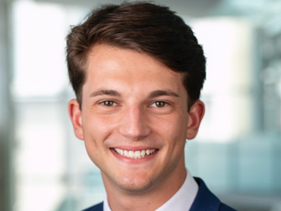 Person in a business suit with a white shirt and tie, photographed in a modern office setting with blurred windows in the background.