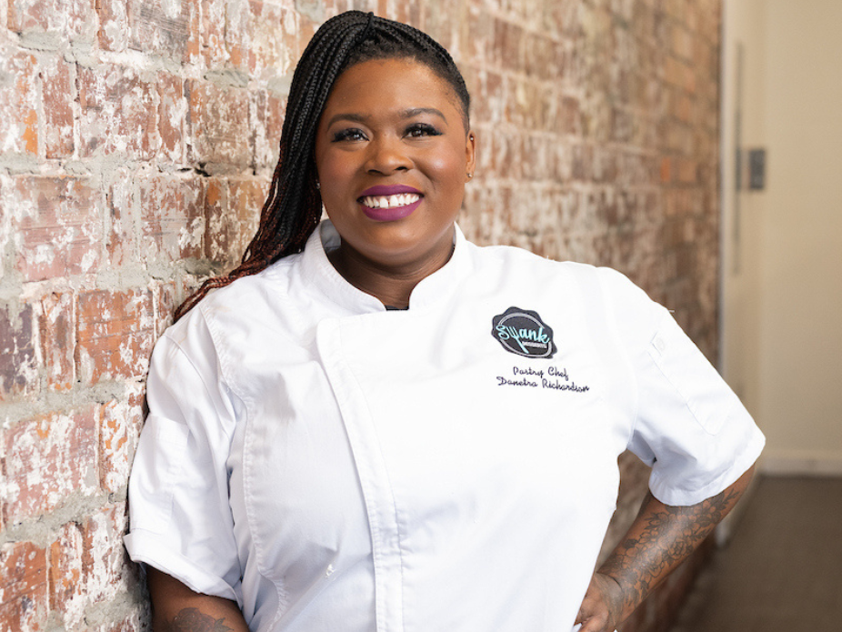 Person in a white chef coat leaning against an exposed brick wall, with embroidered text on the coat indicating a culinary role.