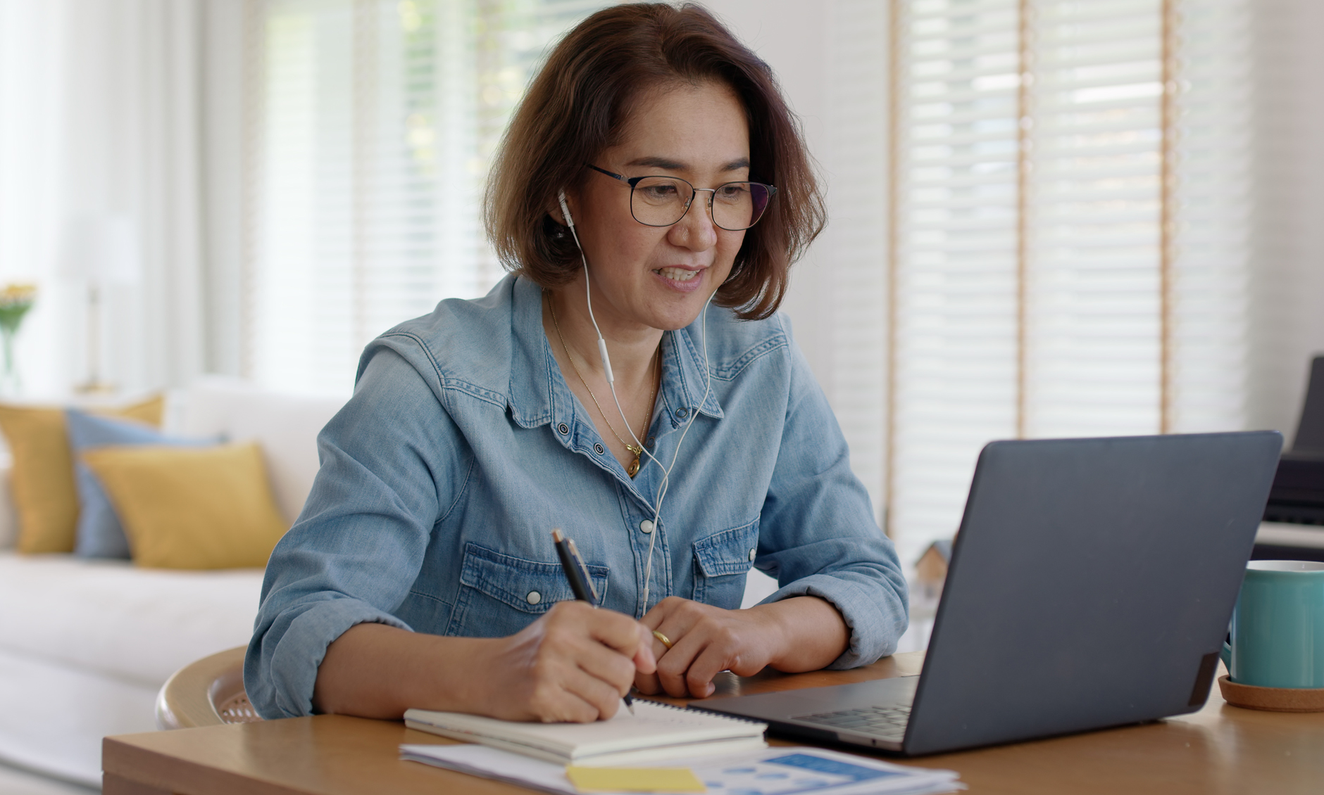 Smiling woman wearing glasses takes notes in a notebook while working on a laptop at a bright home desk, with a coffee mug nearby.