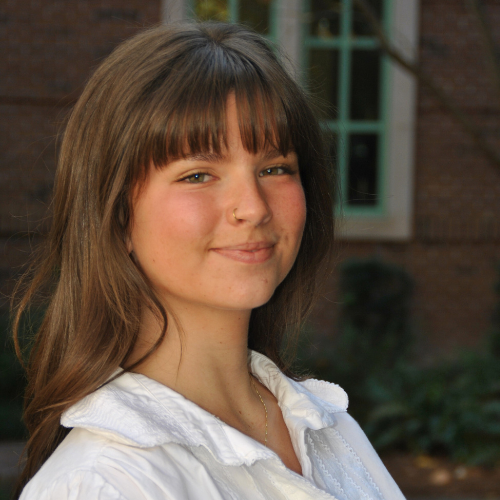 A professional headshot of Della Corvone smiling, wearing a white blouse, in front of an exterior window.