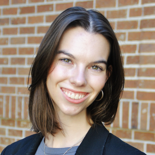 A portrait of Lulu McDowell with short brown hair, wearing a black blazer, smiling and standing in front of a brick exterior.