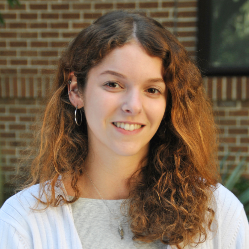 A professional headshot of Maddie Bartlett smiling, wearing a light-colored sweater on a brick background.