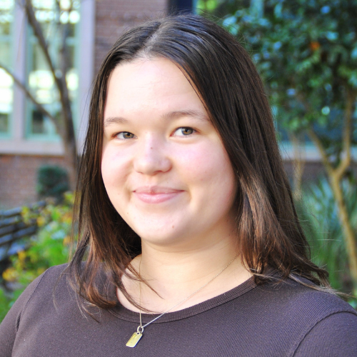 Headshot of a Maddie Johnson with brown hair and wearing a brown sweater and gold necklace, smiling in front of trees.