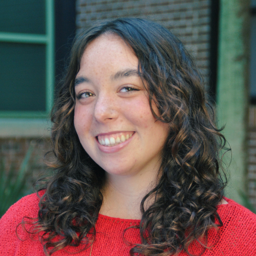 Headshot of Sawyor Plath, with long curly brown hair, smiling for a professional headshot, wearing a red sweater.