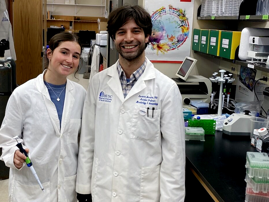 Two researchers in lab coats smiling in a laboratory setting, surrounded by scientific equipment and materials.