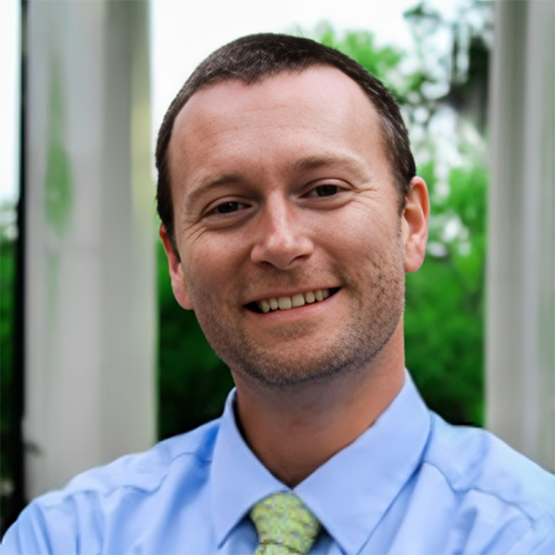 Headshot of Fenner Hall, a smiling man with short hair and stubble beard wearing a blue shirt and green tie.