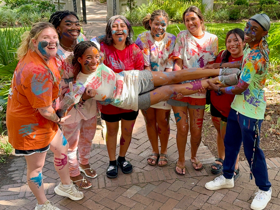 Covered in brightly covered paint, seven SPECTRA participants hold up their group leader while laughing smiling at the camera.