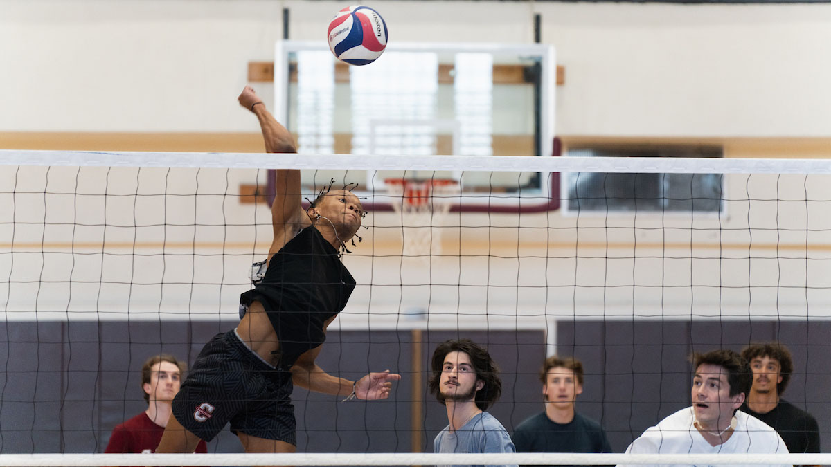 A College of Charleston student jumping to spike a blue, red and white volleyball over a net.