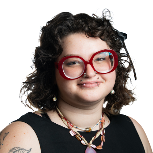 Portrait of Grey Crotts with dark curly hair, bold red glasses, and multiple piercings, wearing a black top and colorful jewelry.